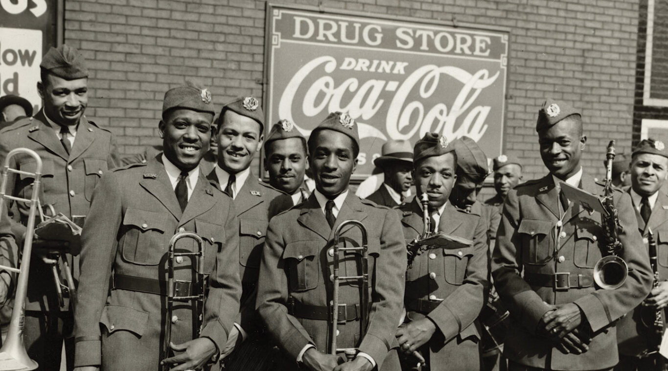 A group of uniformed military band members pose smiling with instruments under vintage Coca-Cola and drugstore signs. The mood is cheerful and proud.