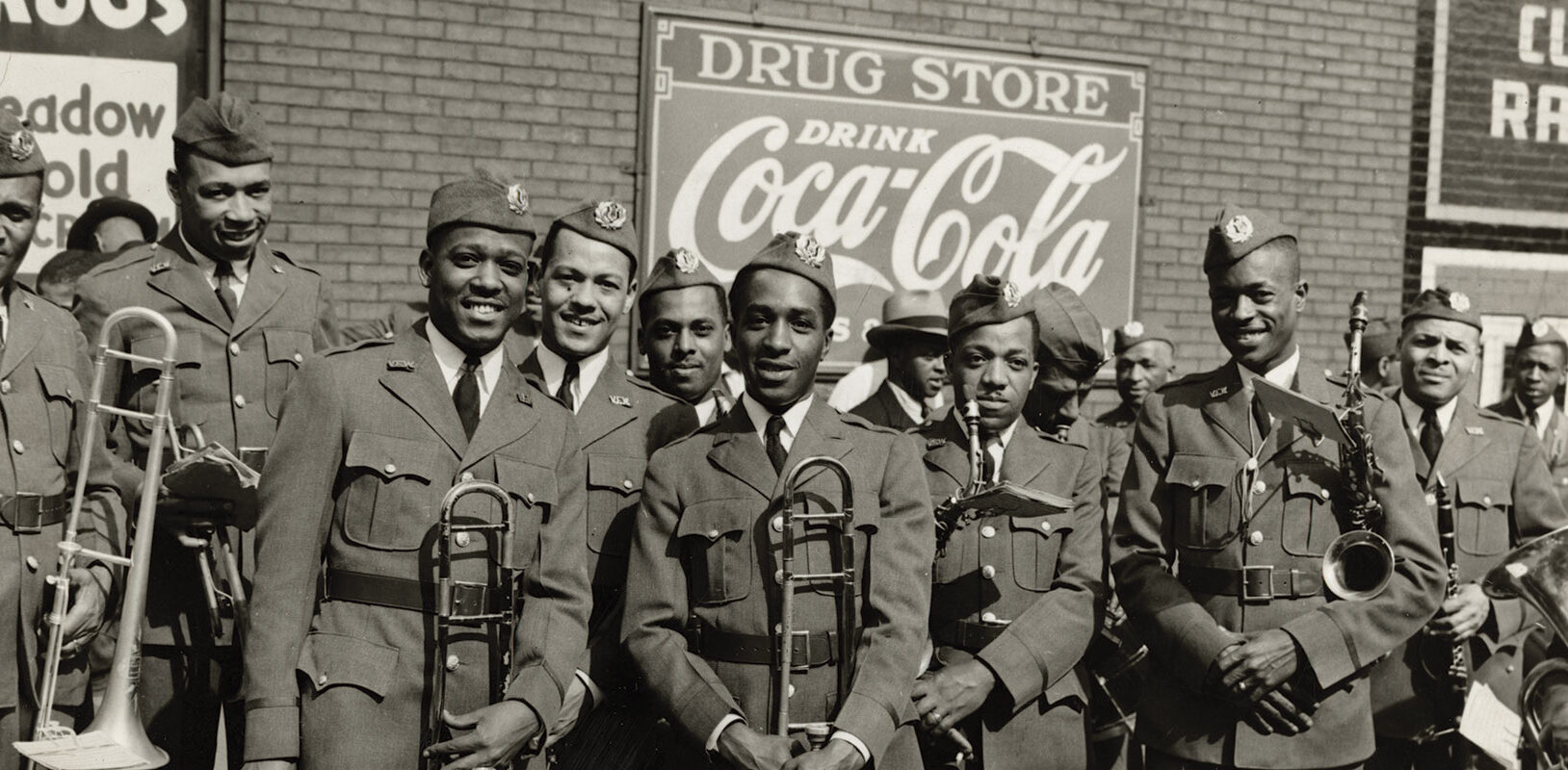 A group of uniformed military band members pose smiling with instruments under vintage Coca-Cola and drugstore signs. The mood is cheerful and proud.