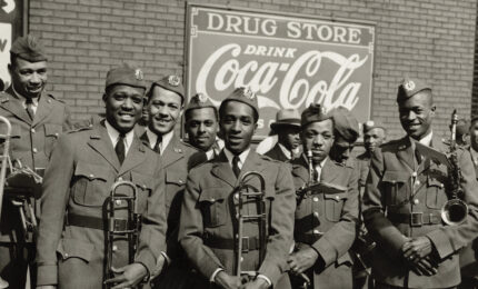 A group of uniformed military band members pose smiling with instruments under vintage Coca-Cola and drugstore signs. The mood is cheerful and proud.