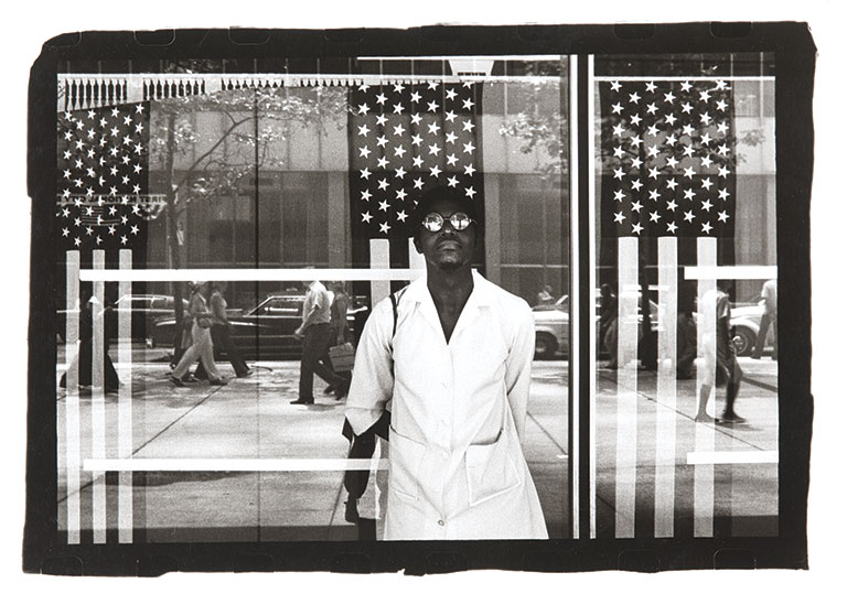 A person in a white coat stands in front of a window adorned with American flag patterns. Reflections of pedestrians and cars are visible, creating a busy urban atmosphere.