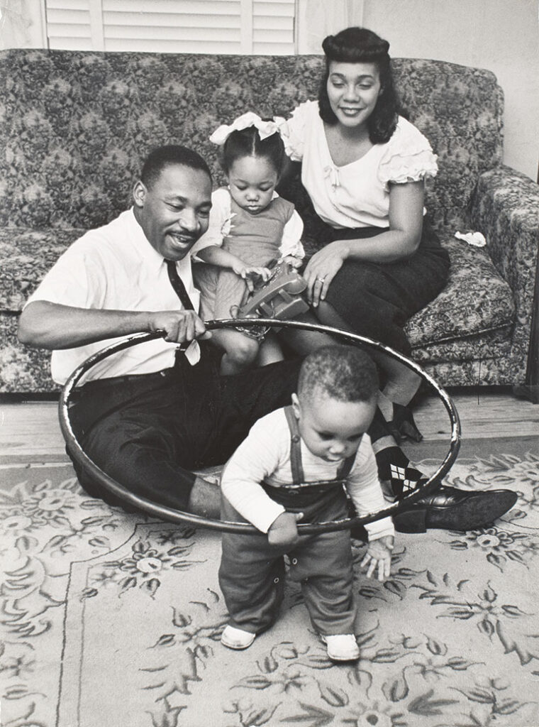 A joyful family scene with a father holding a hula hoop for a toddler to walk through. A mother and another child sit on a floral sofa, smiling warmly.