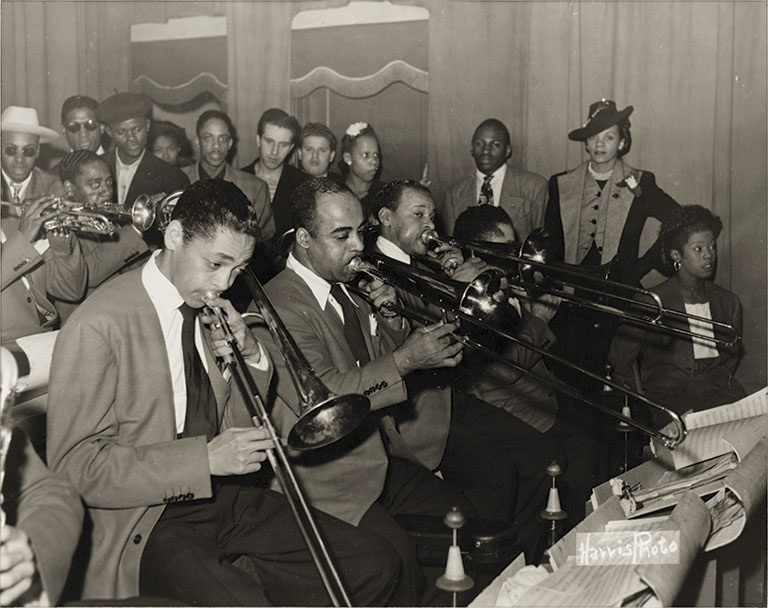 Black-and-white photo of a jazz band playing trombones and trumpets, surrounded by a diverse, well-dressed audience. The scene is lively and engaging.