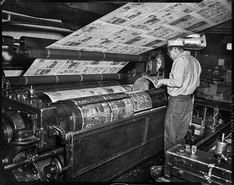 A man in work clothes and cap inspects a large, active newspaper printing press with rotating cylinders and sheets of printed pages. Industrial setting.