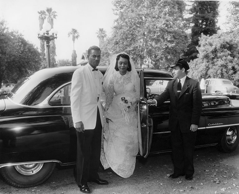 A bride and groom stand beside a classic car. The groom wears a white tuxedo, and the bride is in a lace gown, holding a bouquet. A chauffeur holds the car door open in a park-like setting, exuding an elegant, joyful tone.