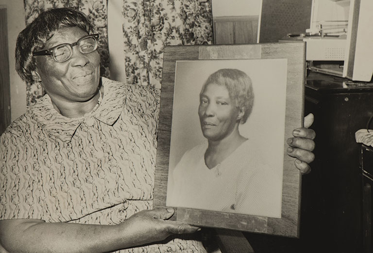An elderly woman with glasses holds a framed portrait of another woman. She smiles warmly, suggesting fond remembrance. Floral curtains in the background.