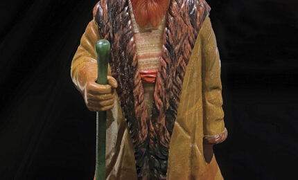 A detailed statuette of a bearded man in a long, fur-trimmed coat, holding a green walking stick. He wears a hat, exuding a rustic, thoughtful presence.