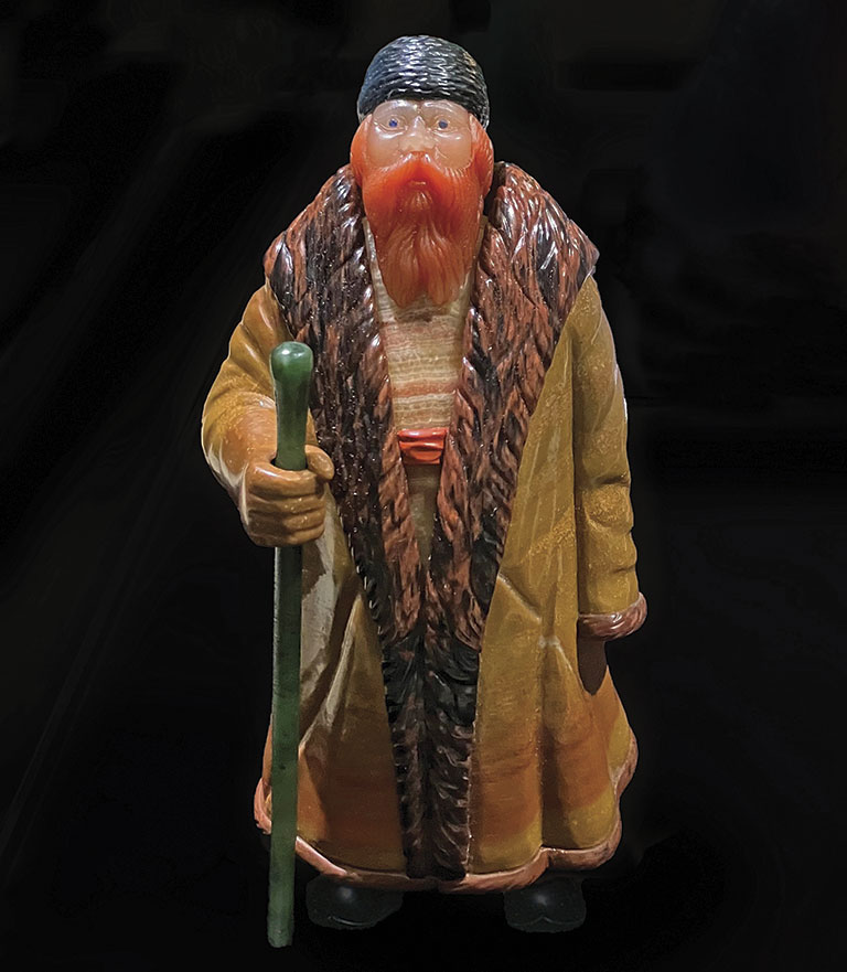 A detailed statuette of a bearded man in a long, fur-trimmed coat, holding a green walking stick. He wears a hat, exuding a rustic, thoughtful presence.