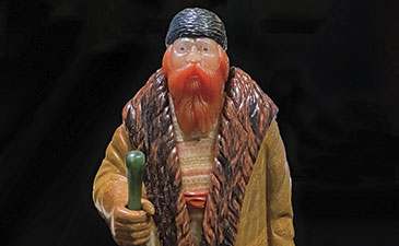 A detailed statuette of a bearded man in a long, fur-trimmed coat, holding a green walking stick. He wears a hat, exuding a rustic, thoughtful presence.