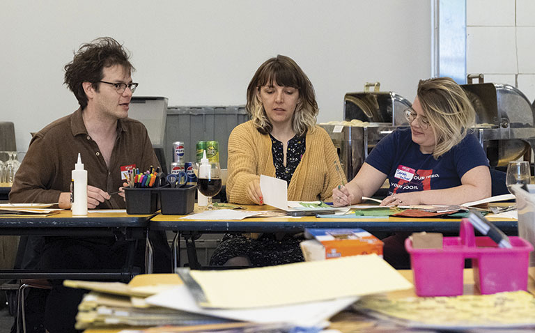 Three people sit around a cluttered table, engaging in a creative activity. Supplies and papers are scattered, creating a collaborative and focused atmosphere.
