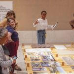 A diverse group of people stands around a table with architectural models and photos. A person speaks enthusiastically, engaging the attentive group.