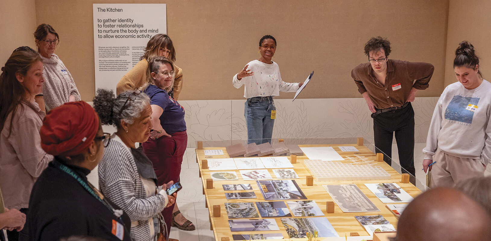 A diverse group of people stands around a table with architectural models and photos. A person speaks enthusiastically, engaging the attentive group.