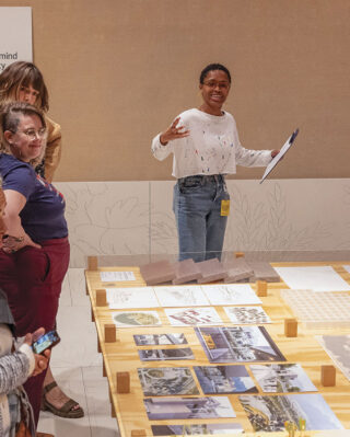 A diverse group of people stands around a table with architectural models and photos. A person speaks enthusiastically, engaging the attentive group.
