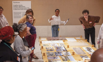 A diverse group of people stands around a table with architectural models and photos. A person speaks enthusiastically, engaging the attentive group.