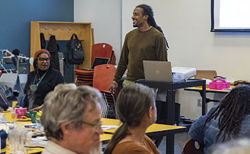 A speaker presents at a workshop or meeting, with attendees seated at tables, engaged in discussion and activities.