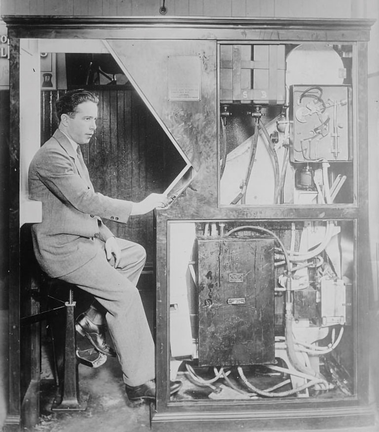 A man in a suit sits inside a vintage mechanical booth, intently reading a document. The booth is filled with complex machinery, evoking a sense of curiosity.