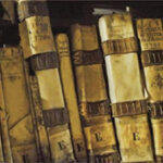 A row of aged, leather-bound books with faded, handwritten titles and ornate designs on their spines. The scene conveys a sense of history and antiquity.