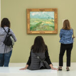 People in a museum admire landscape paintings. A central figure sits, viewing a green field painting, while a family takes a photo. The scene is lively and engaged.