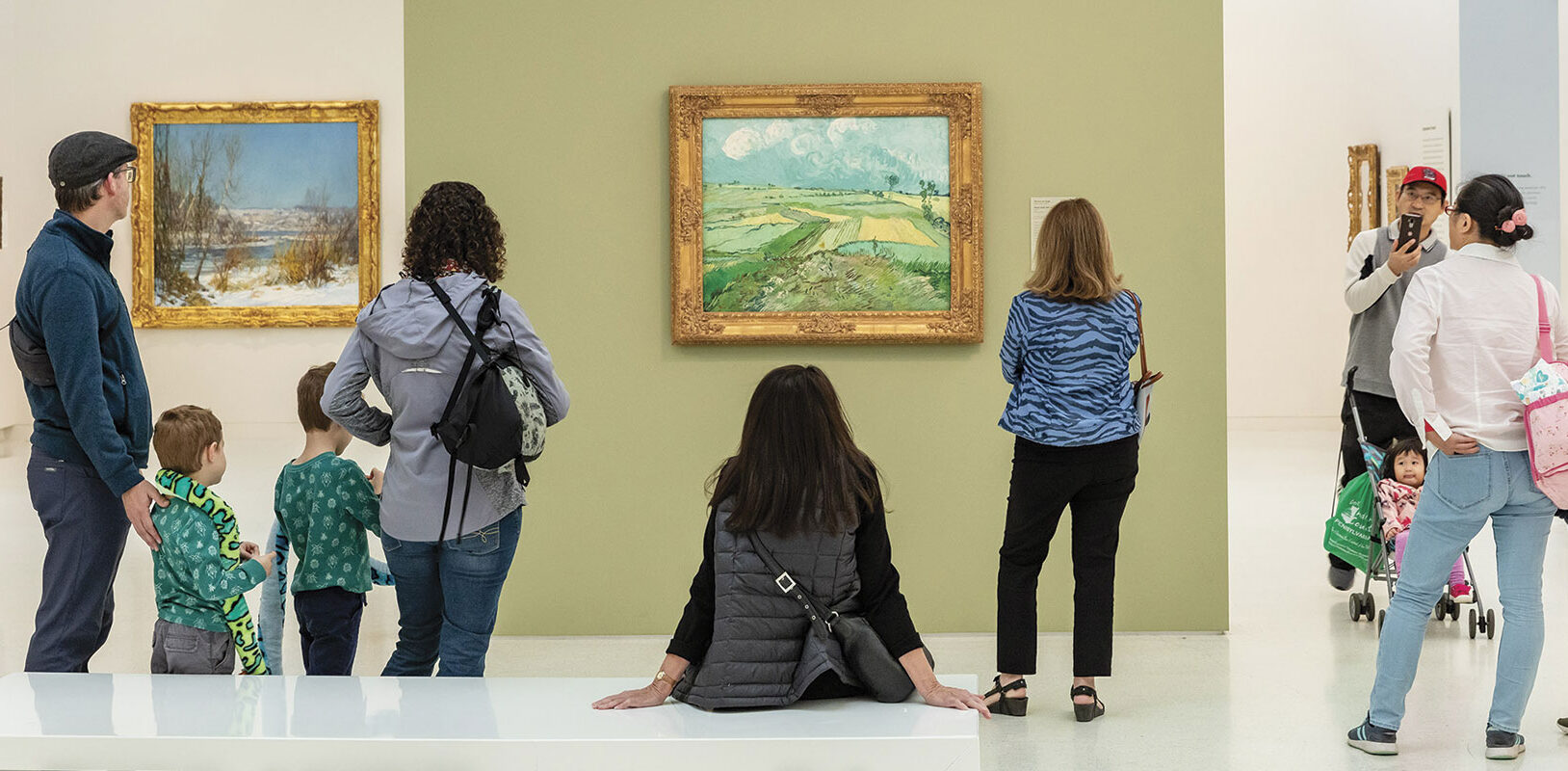People in a museum admire landscape paintings. A central figure sits, viewing a green field painting, while a family takes a photo. The scene is lively and engaged.
