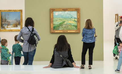 People in a museum admire landscape paintings. A central figure sits, viewing a green field painting, while a family takes a photo. The scene is lively and engaged.