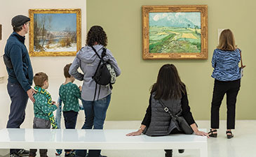 People in a museum admire landscape paintings. A central figure sits, viewing a green field painting, while a family takes a photo. The scene is lively and engaged.