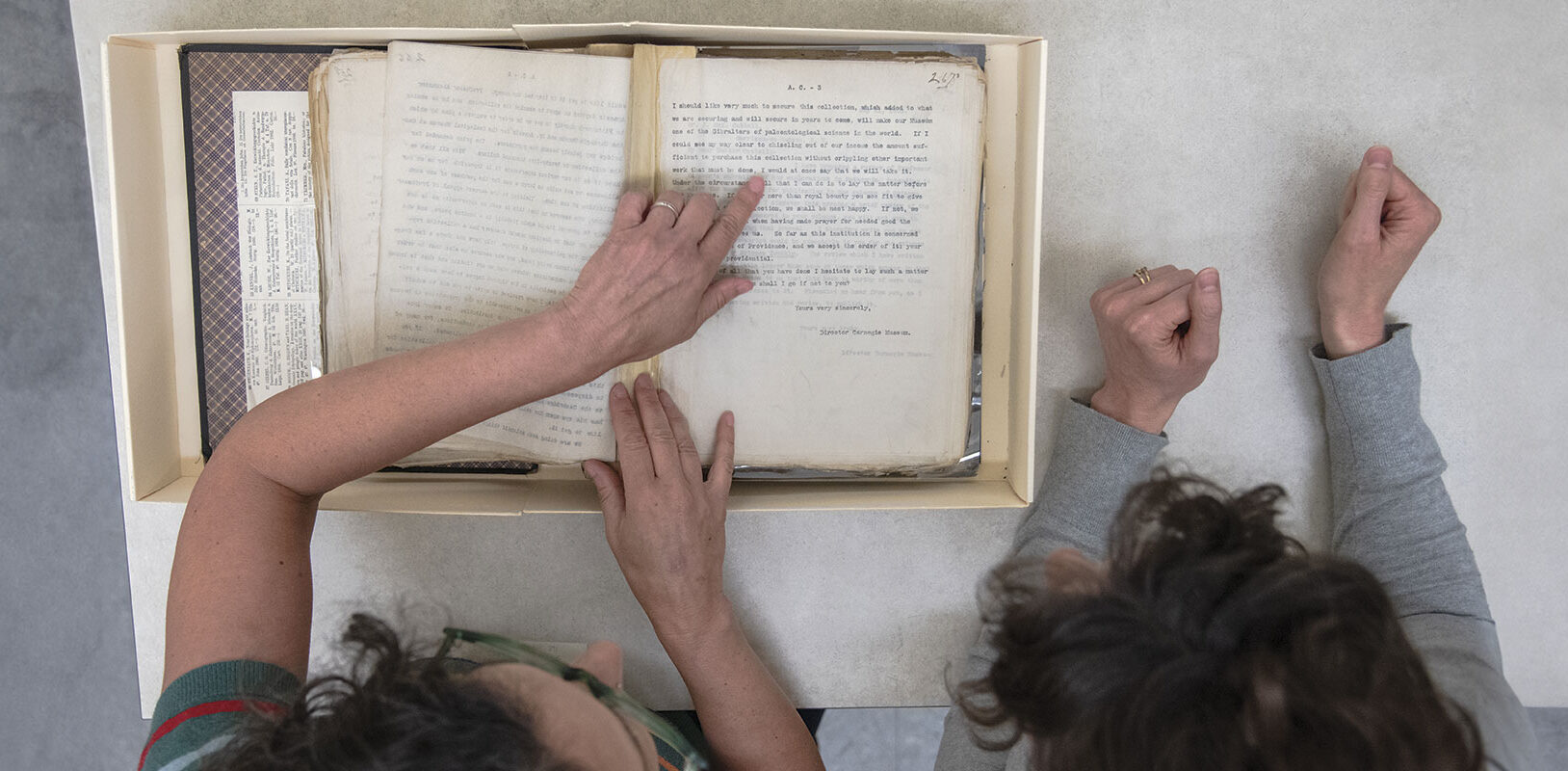 Two people examine an open archival box with historical documents, pointing and discussing the contents together.