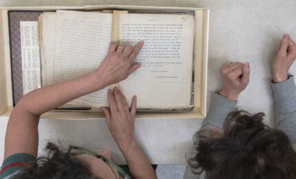 Two people examine an open archival box with historical documents, pointing and discussing the contents together.