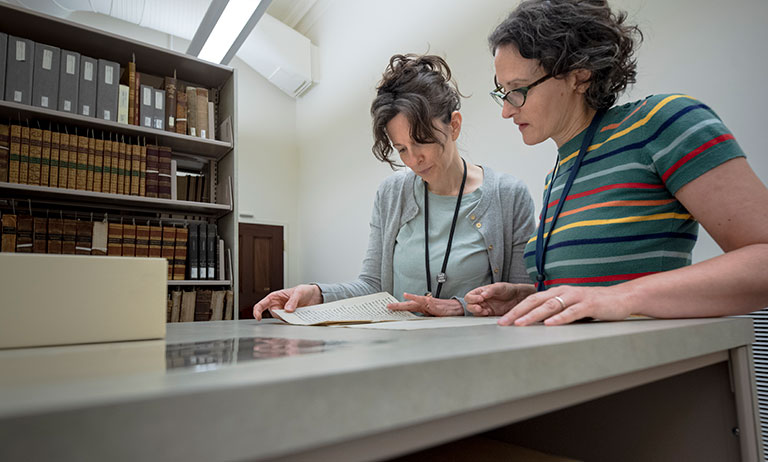 Two individuals examine an open book on a table, surrounded by shelves filled with various books and archival materials.