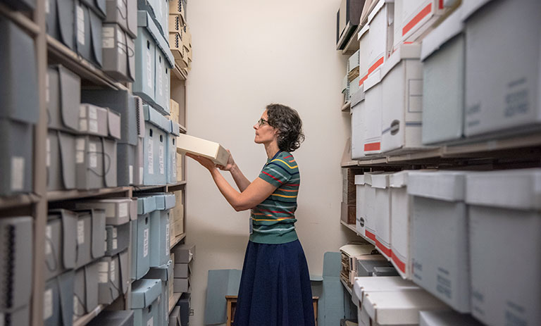 A person examines a document inside a narrow archive room filled with organized boxes on both sides.