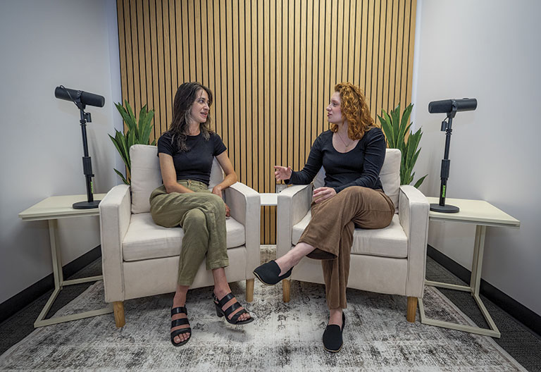 Two women sit in armchairs having a conversation in a modern studio. Microphones on stands flank the chairs, and plants add natural decor.