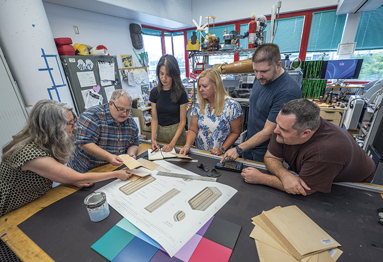 Six people collaborate around a table with design plans and material samples in a creative workspace. The atmosphere is focused and cooperative.