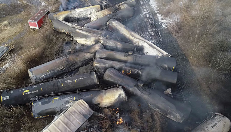 Aerial view of a train derailment shows overturned black tanker cars scattered across tracks, smoke rising, with nearby trees and a red truck. Somber scene.