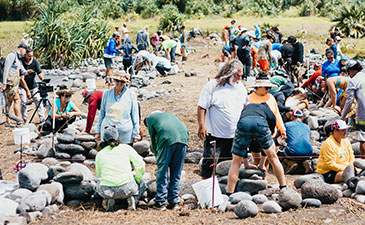 A group of people work together outdoors, carefully arranging stones on the ground under a sunny sky, amidst lush greenery.
