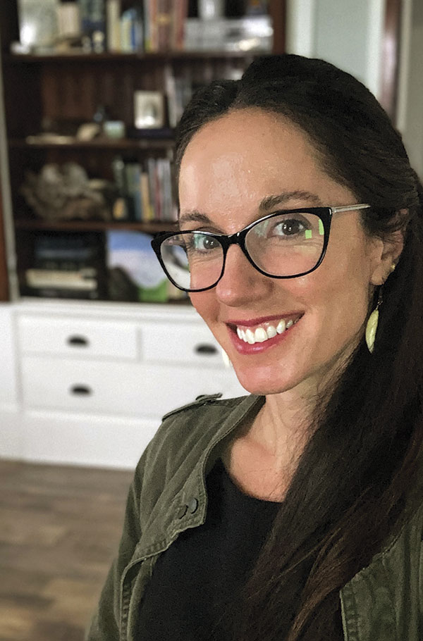 A woman with long dark hair and glasses smiles warmly in an indoor setting. She wears a green jacket. Shelves with books and decor are blurred in the background.