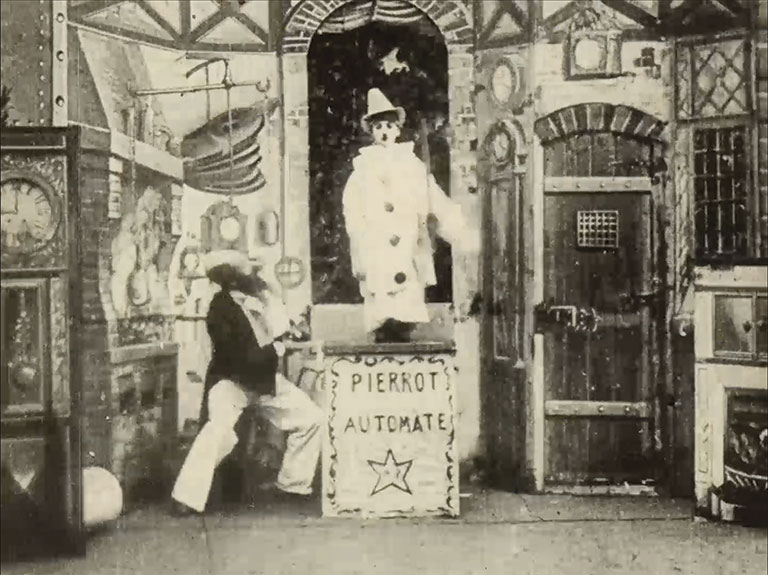 Vintage shop display with a seated figure and white Pierrot mannequin behind a box labeled “Pierrot Automate,” surrounded by clocks and antiques.