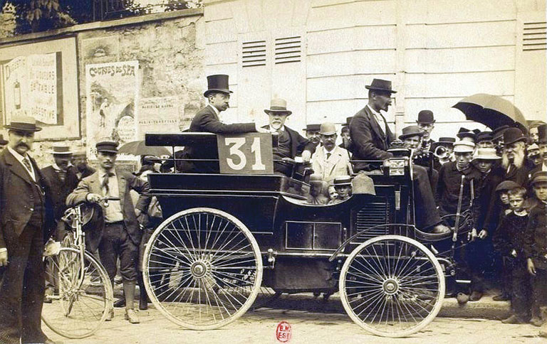 Sepia historic photograph of an early race car marked 31, surrounded by formally dressed men, cyclists, and a crowd with umbrellas.