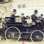 Sepia historic photograph of an early race car marked 31, surrounded by formally dressed men, cyclists, and a crowd with umbrellas.