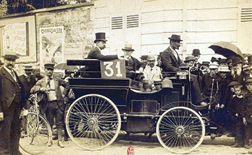 Sepia historic photograph of an early race car marked 31, surrounded by formally dressed men, cyclists, and a crowd with umbrellas.