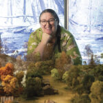 Smiling woman in glasses leans over a miniature forest landscape, with autumn-colored trees and a snowy woodland scene behind her.