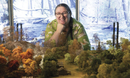 Smiling woman in glasses leans over a miniature forest landscape, with autumn-colored trees and a snowy woodland scene behind her.
