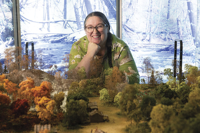 Smiling woman in glasses leans over a miniature forest landscape, with autumn-colored trees and a snowy woodland scene behind her.