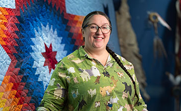 Smiling woman with glasses and braided hair poses in a green animal-print shirt before a colorful red, blue, and white quilt.