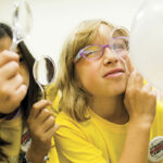 Friends playfully examine a large white balloon with magnifying glasses; a smiling woman in yellow glasses and shirt appears curious.