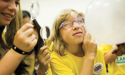 Friends playfully examine a large white balloon with magnifying glasses; a smiling woman in yellow glasses and shirt appears curious.