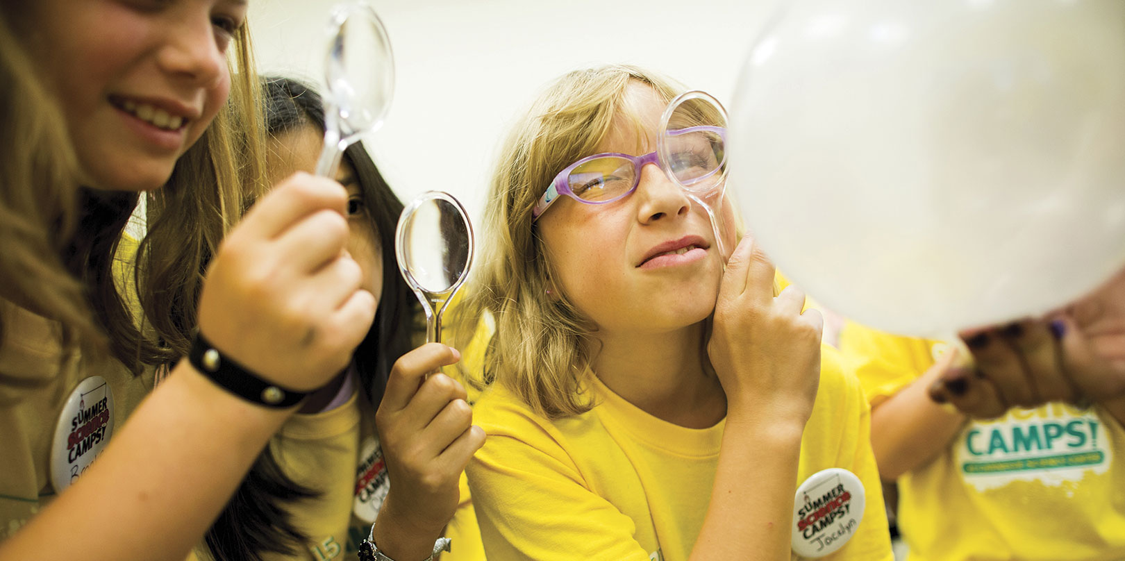 Friends playfully examine a large white balloon with magnifying glasses; a smiling woman in yellow glasses and shirt appears curious.