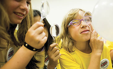Friends playfully examine a large white balloon with magnifying glasses; a smiling woman in yellow glasses and shirt appears curious.