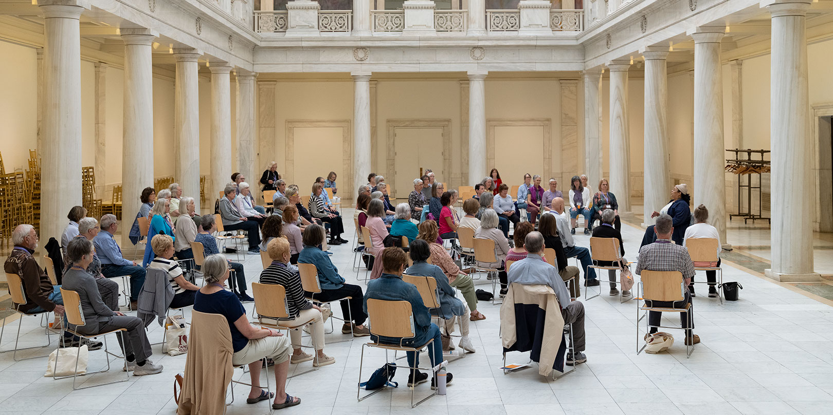 A diverse audience sits in a circle for a discussion inside a grand neoclassical hall, surrounded by columns, balconies, and marble floors.