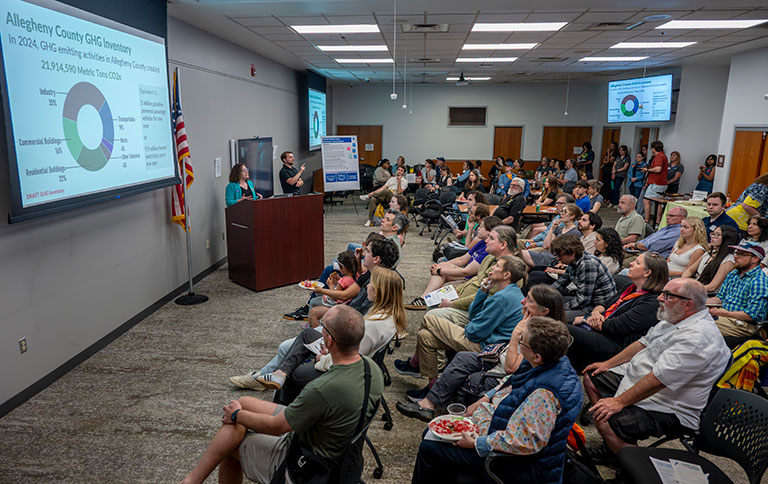 A crowded community meeting features a speaker at a podium, a projected health-data chart, and an attentive audience in a lecture room.
