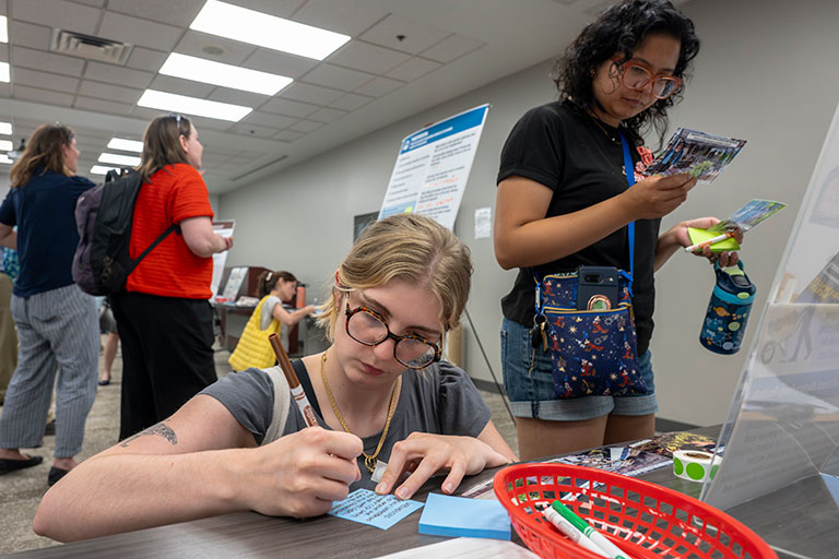 Busy registration table: a woman with glasses fills out a form while another checks brochures; visitors wait behind in a welcoming setting.