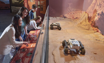 Museum visitors explore an interactive Mars exhibit, viewing robotic rovers on a red, rocky landscape through glass; curious mood.