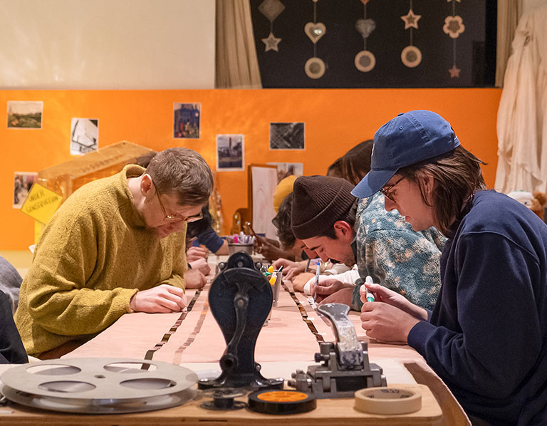 People collaborate at a printmaking workshop, carefully crafting designs at a long table in a warm, creative studio with colorful artwork.